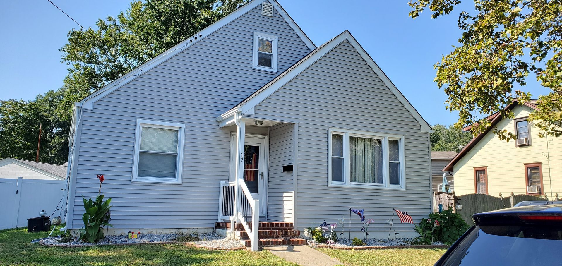 Small light-gray house with white trim, front porch, and two cars parked outside on a sunny day