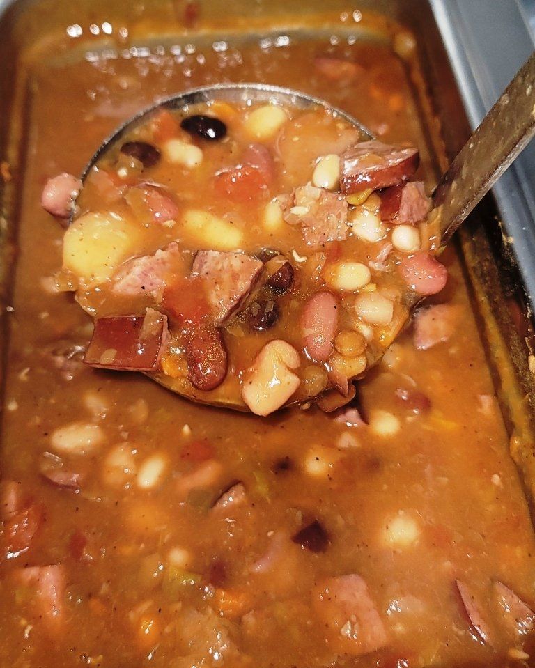 A ladle scoops a thick bean soup with sausage and vegetables from a serving tray.