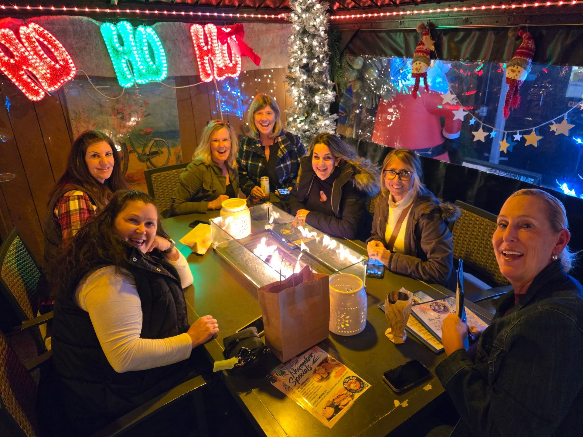 Group of women smiling around a table with a fire pit, festive decorations in a lit outdoor setting.