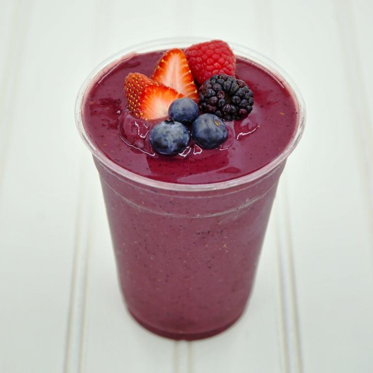 Berry smoothie in a clear plastic cup, topped with fresh berries on a white wood surface.