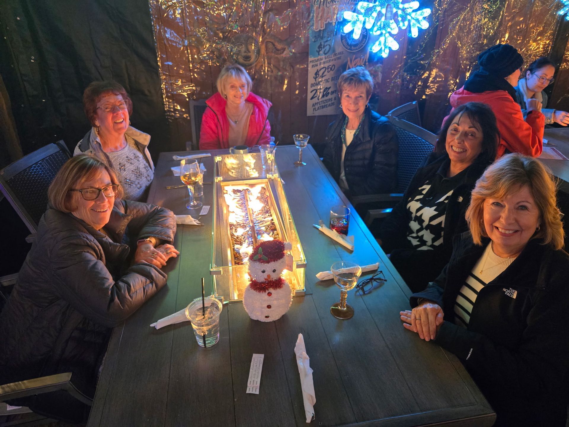 Group of people smiling around an outdoor table with a fire feature and a snowman.
