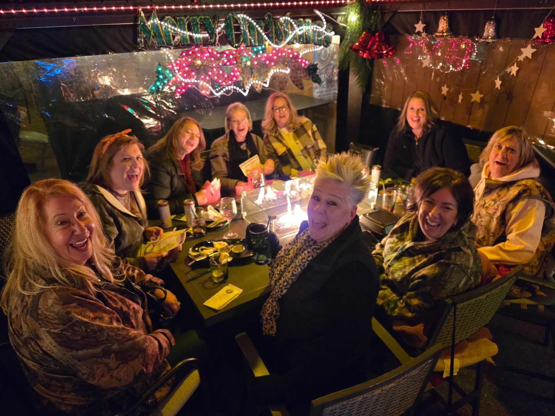 A group of people smiles around a lit outdoor table, decorated with lights, at night.