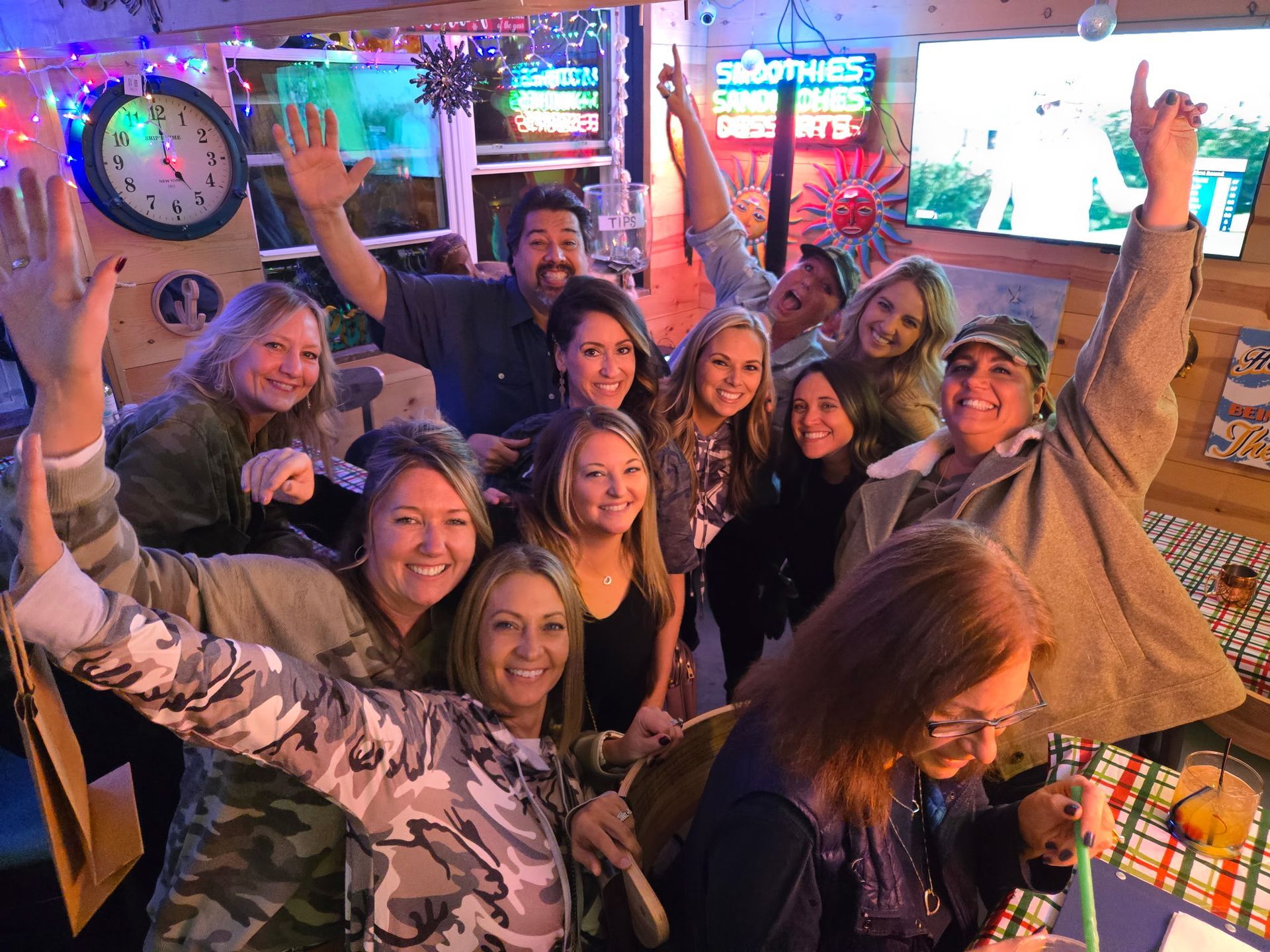 Group of people smiling, raising arms in a brightly lit room with a clock and neon signs.