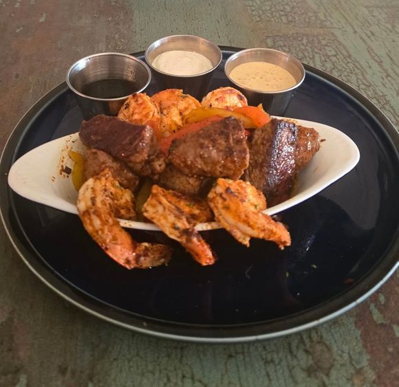 A white oval bowl of grilled shrimp and steak bites with three small metal cups of dipping sauces on a dark plate.