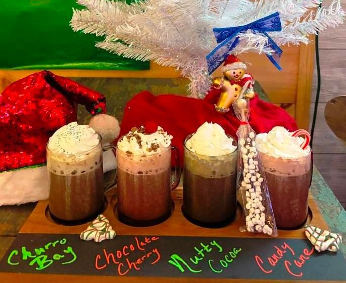 Four holiday-themed hot chocolates on a wooden tray, decorated with whipped cream, Christmas treats, and a Santa hat.