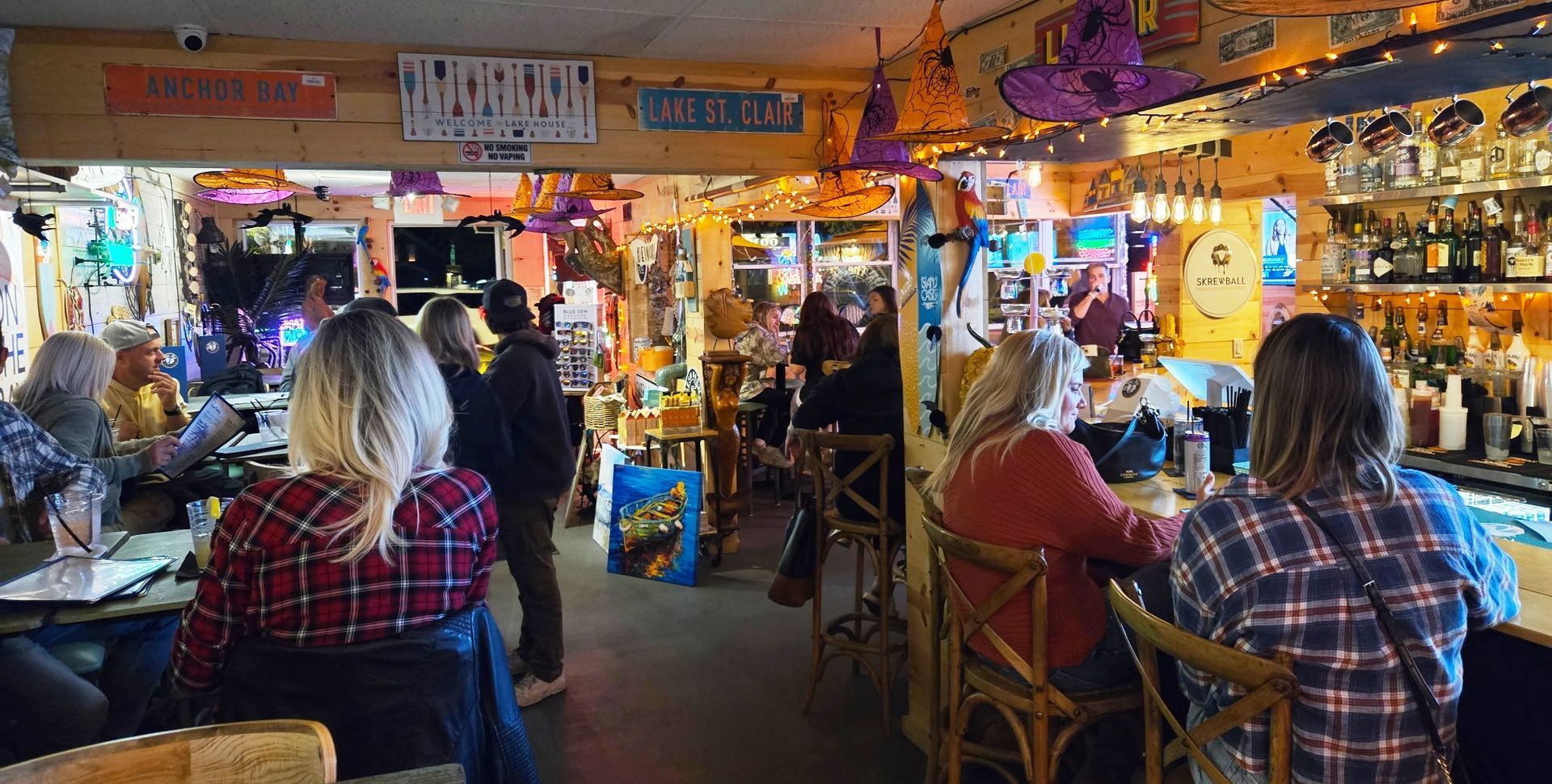 People at a lively, decorated bar, some seated at the counter, others at tables. Yellow and orange tones.