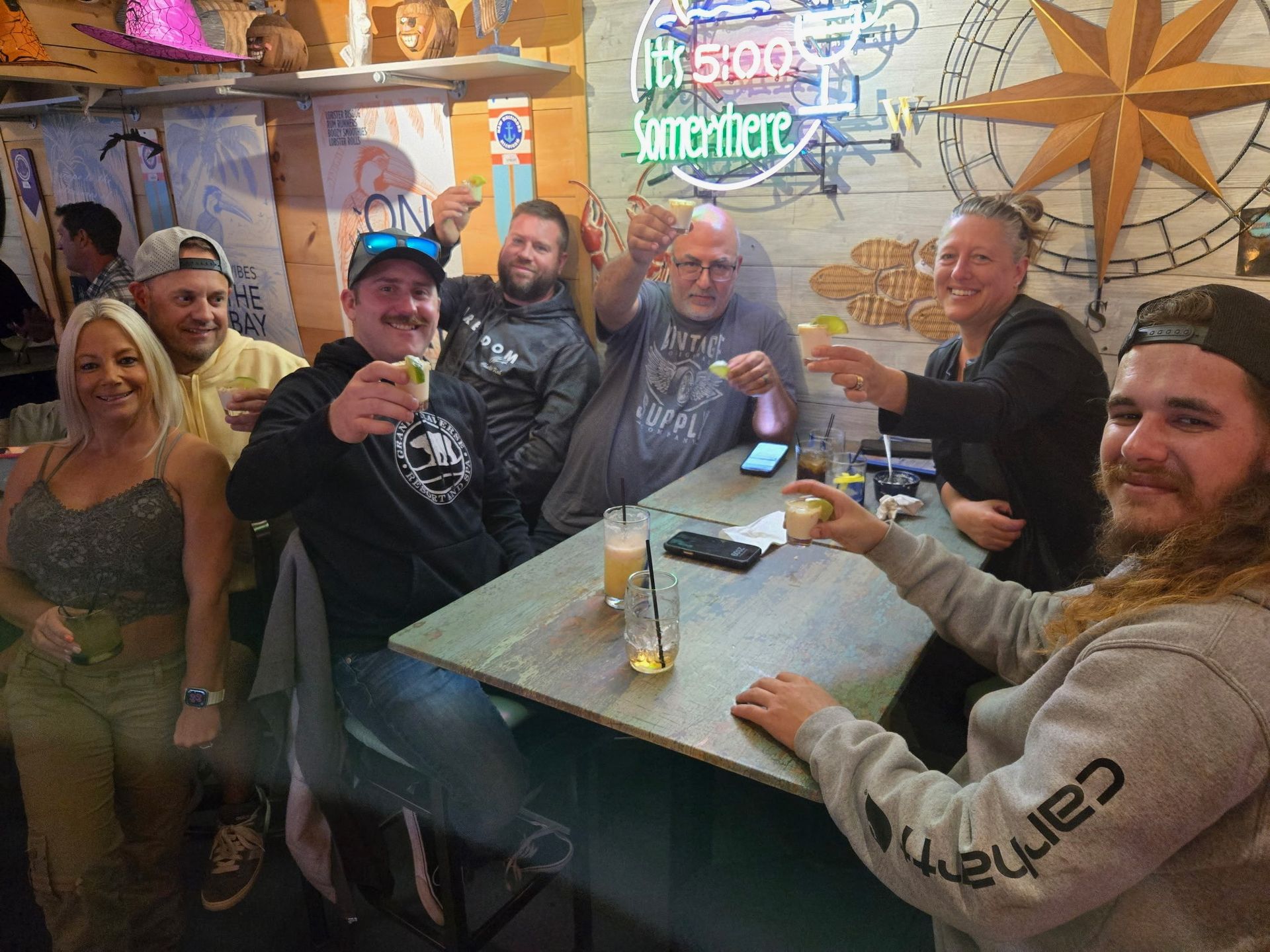 Group of people toasting drinks at a bar. Men and women smiling. Wooden decor and neon sign.