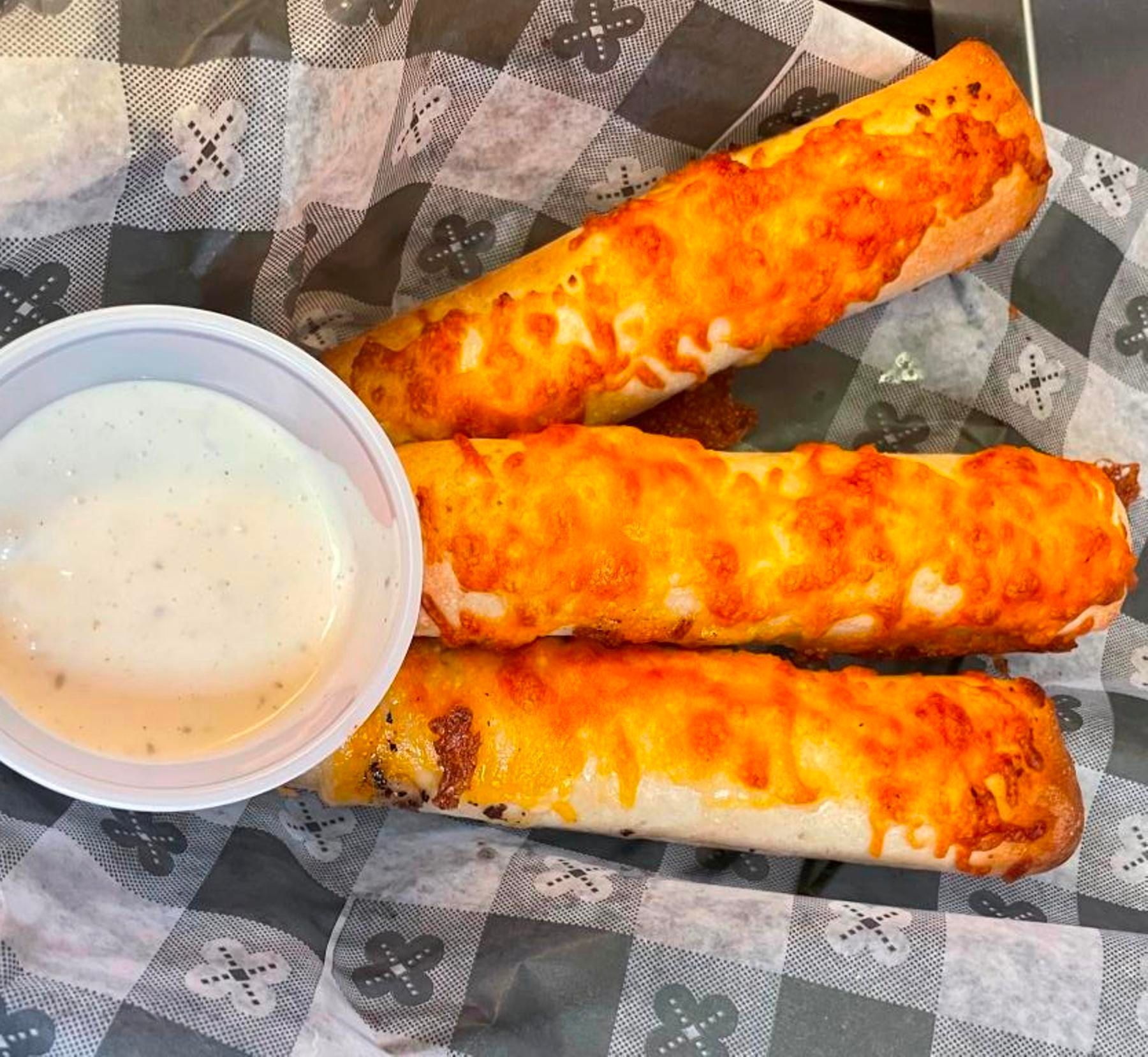 Three cheesy breadsticks with dipping sauce on checkered paper.