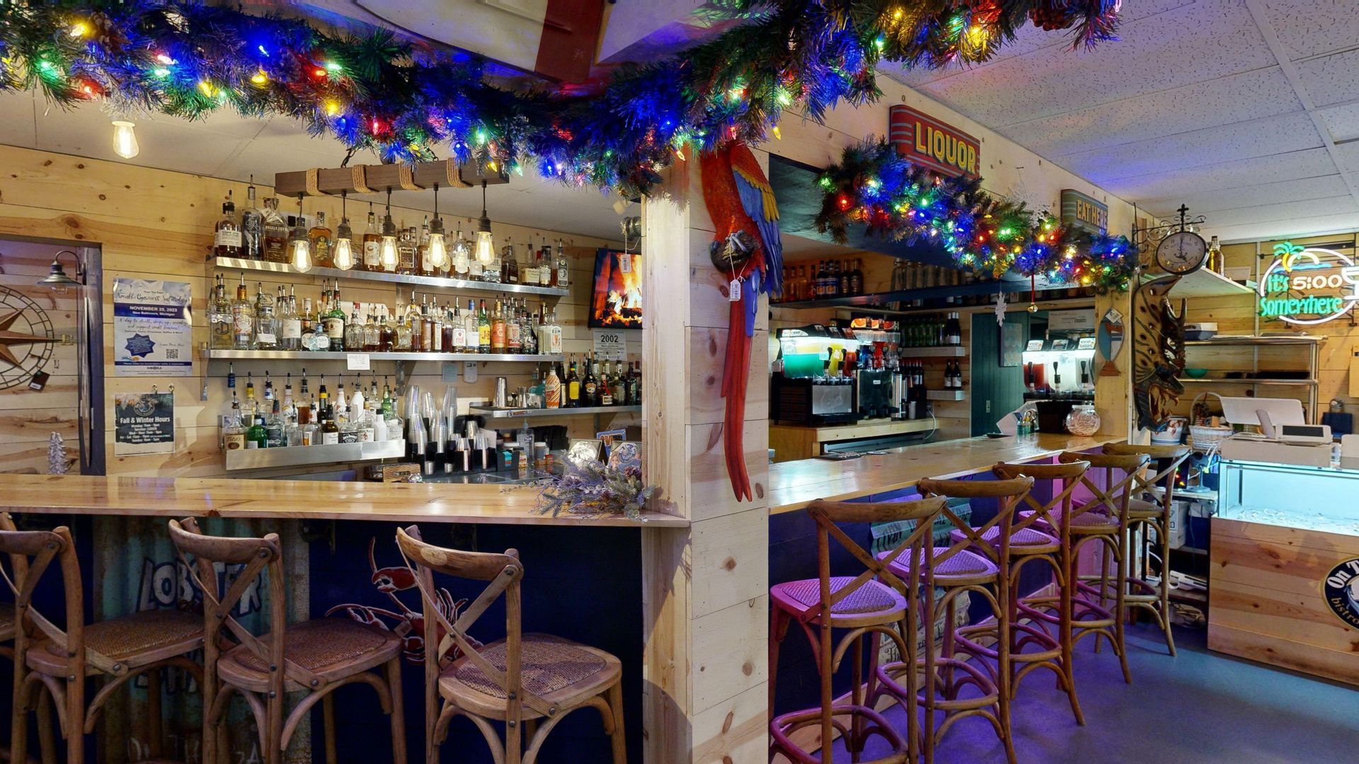 Bar interior with Christmas decorations; wooden bar, stools, liquor bottles, and festive lights.