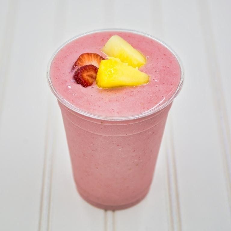 Strawberry-pineapple smoothie in a clear cup, garnished with fruit, on a white-lined surface.