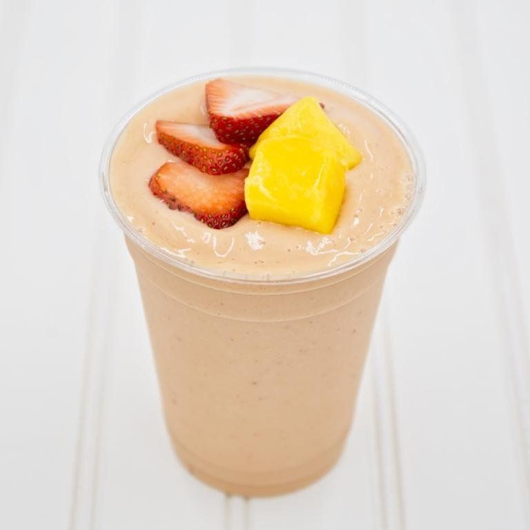 Smoothie in a clear cup, topped with sliced strawberries and mango, on a white surface.