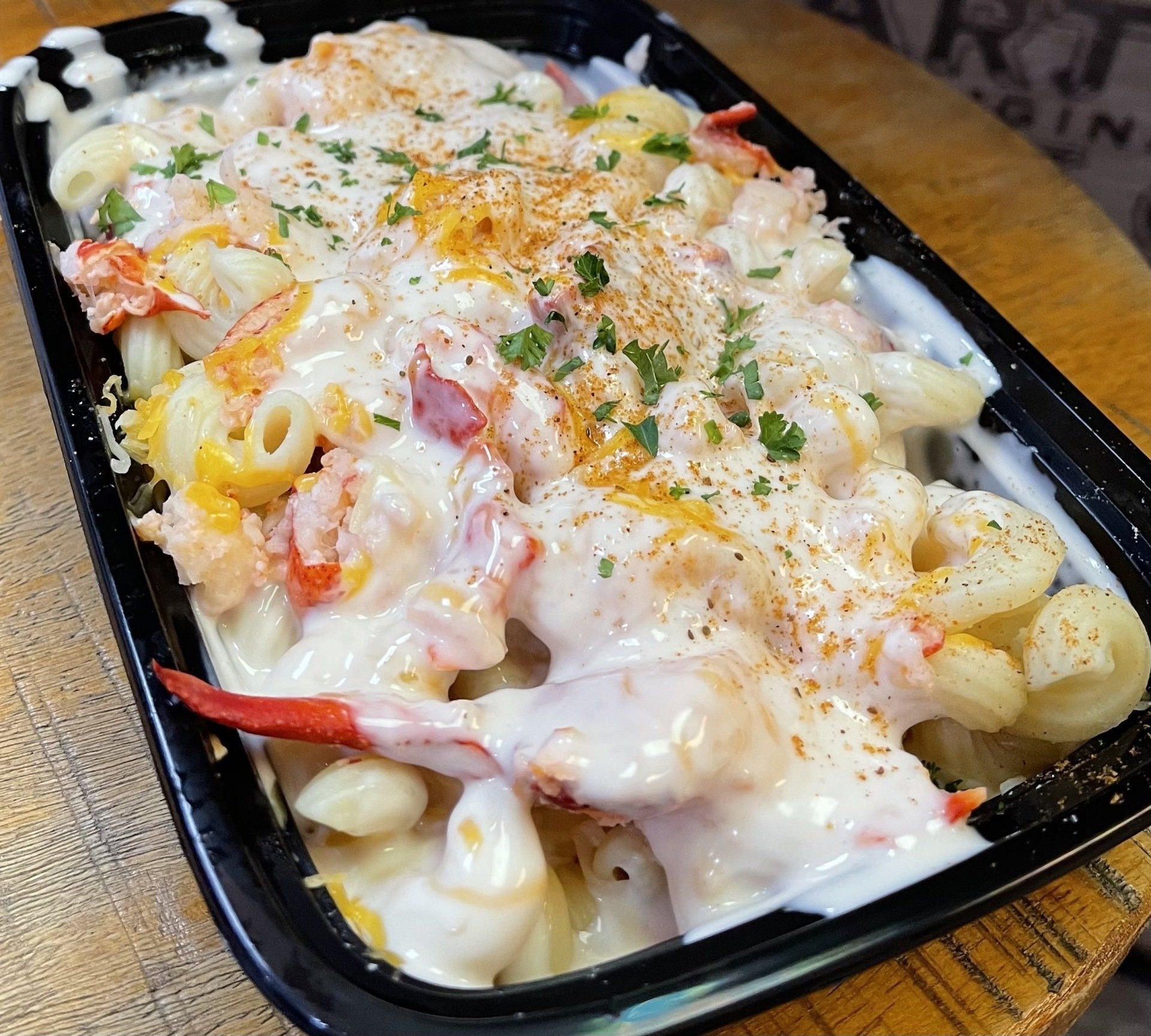 Lobster mac and cheese in a black container; creamy white sauce, lobster claws, sprinkled with parsley.