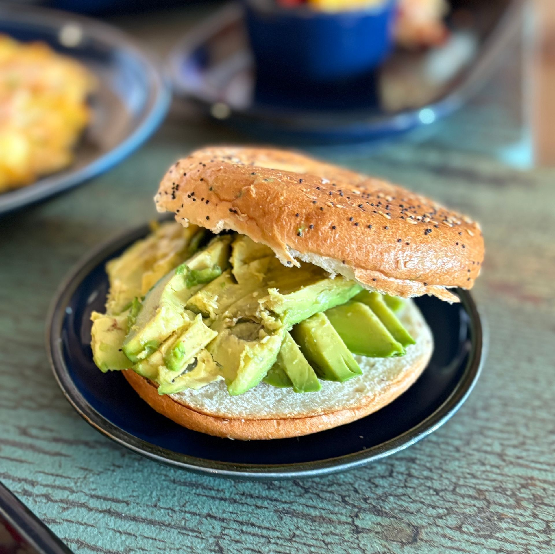 Avocado slices on a bagel with poppy seeds, on a blue plate, on a wooden table.
