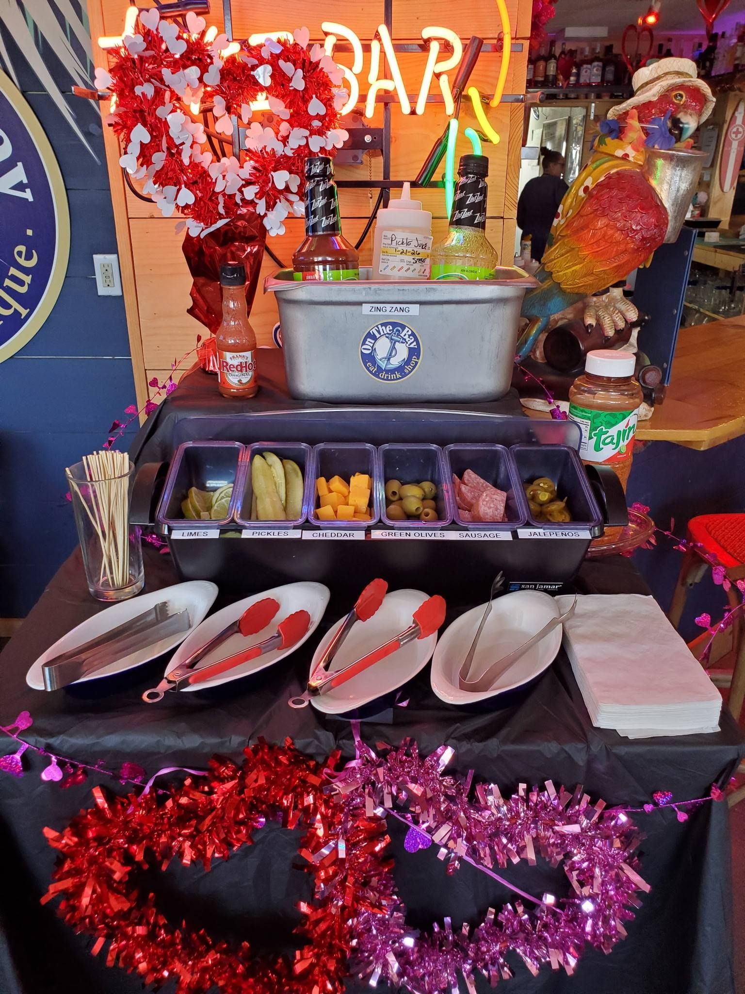 A bloody mary bar with various toppings and serving utensils, under a neon 