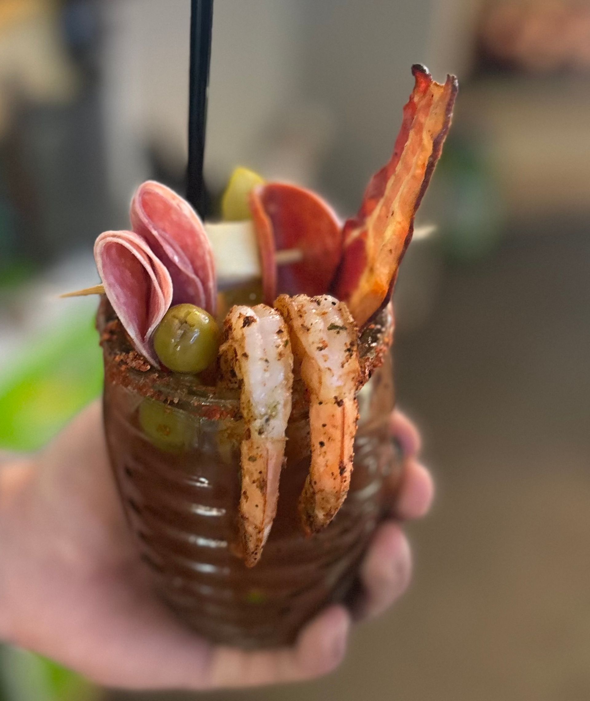 Bloody Mary cocktail in a brown glass, garnished with shrimp, bacon, meats, and olives.