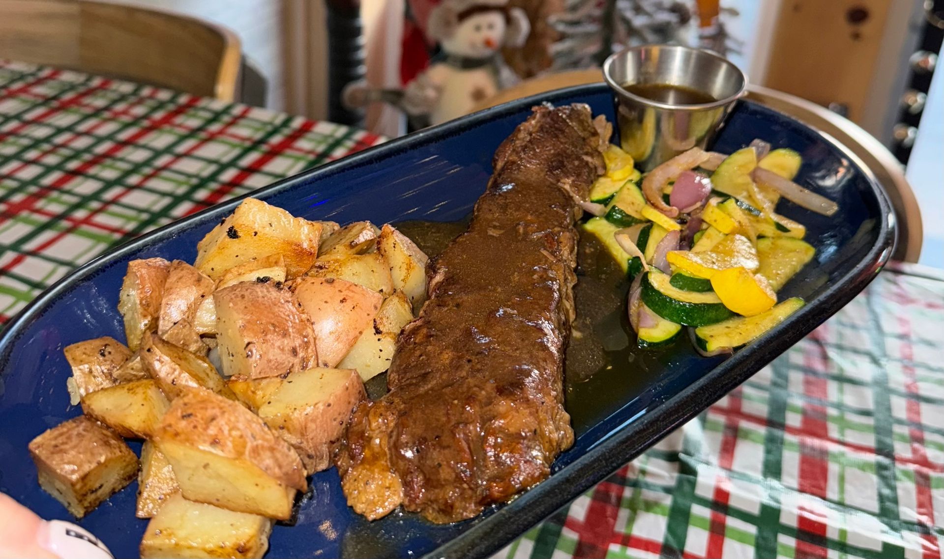 Black platter with grilled steak, roasted potatoes, and mixed vegetables on a checkered tablecloth