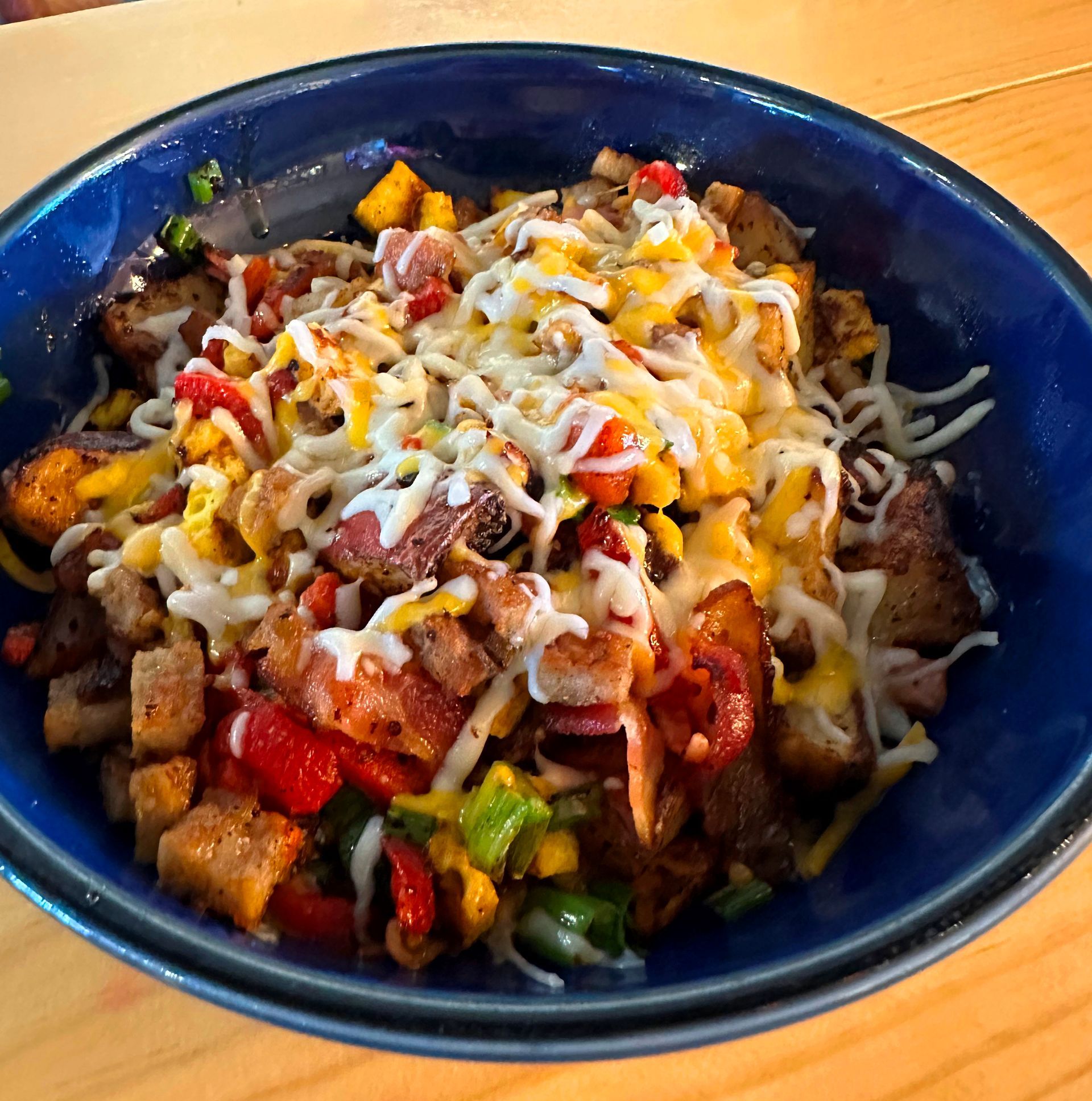 Bowl of breakfast hash: potatoes, peppers, bacon, cheese, green onions in a blue bowl.