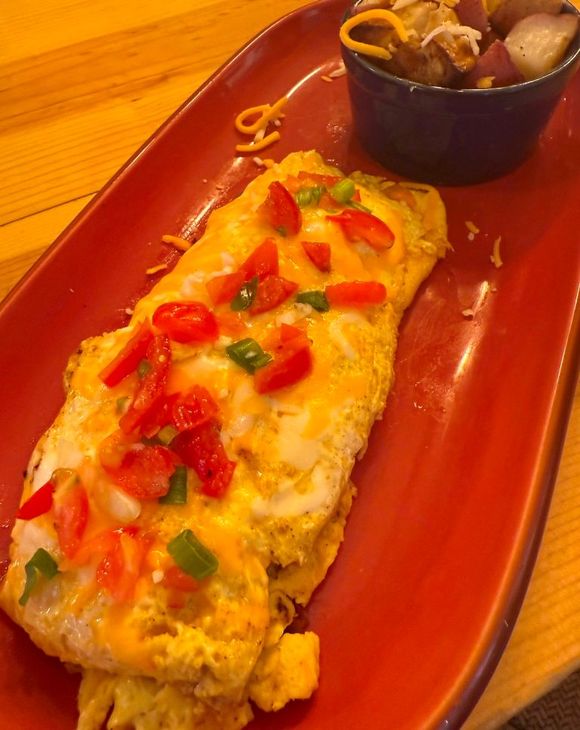 Omelet topped with tomatoes, green onions, and cheese on a red plate, served with a side of potatoes.