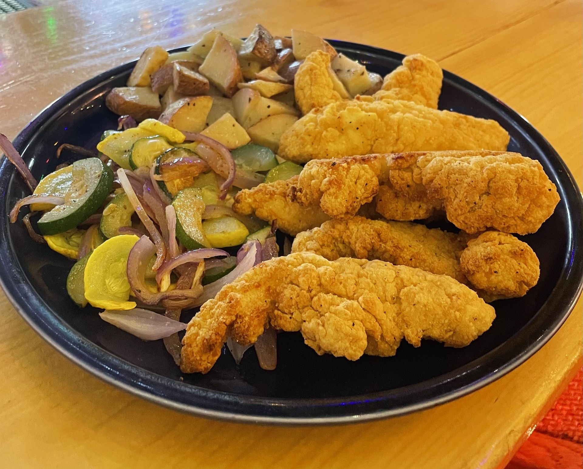 Fried fish fillets with potato wedges and a lemon cucumber salad on a black plate.