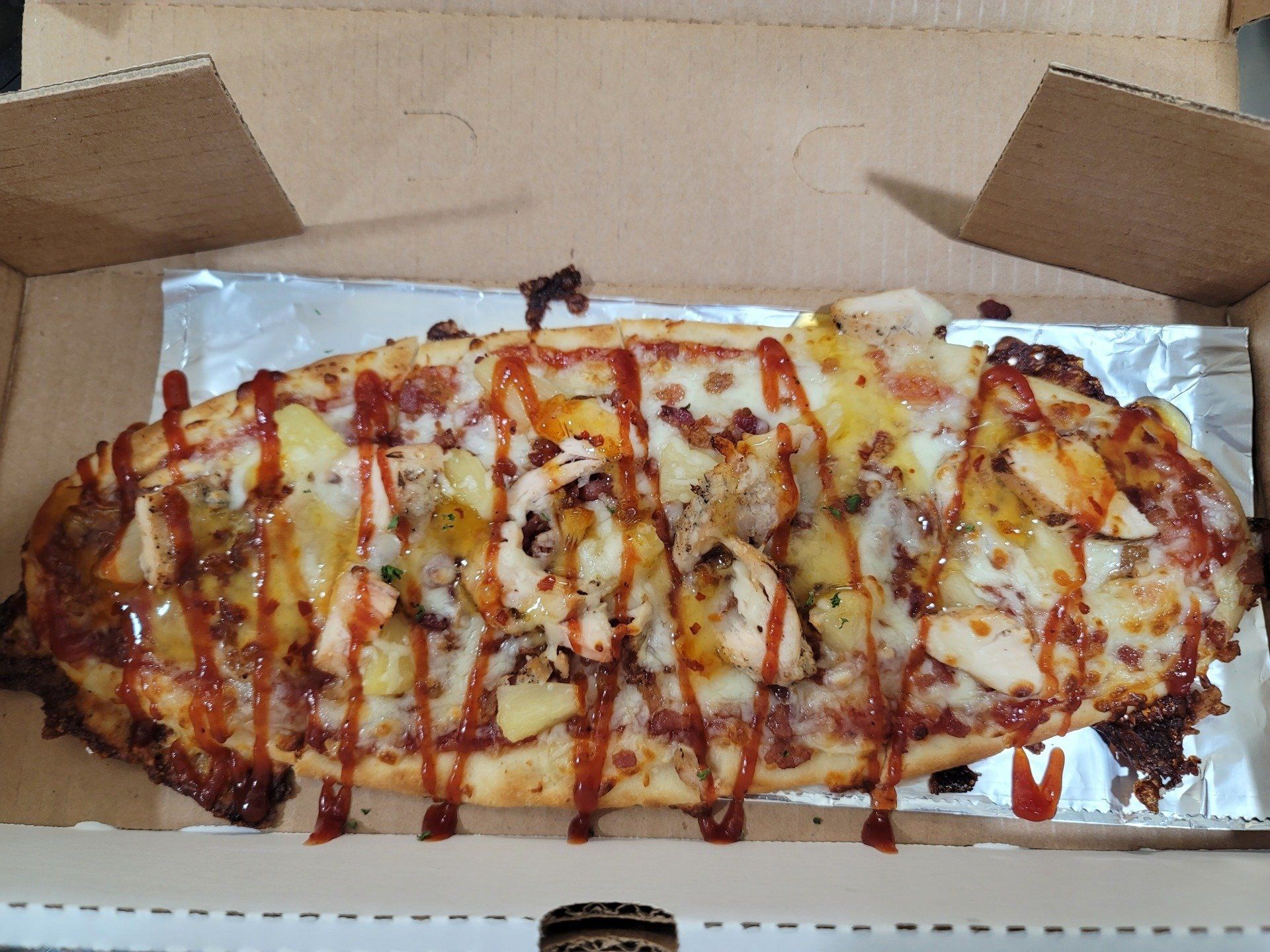 Pizza in a box, topped with chicken, pineapple, bacon, and drizzled with sauce.