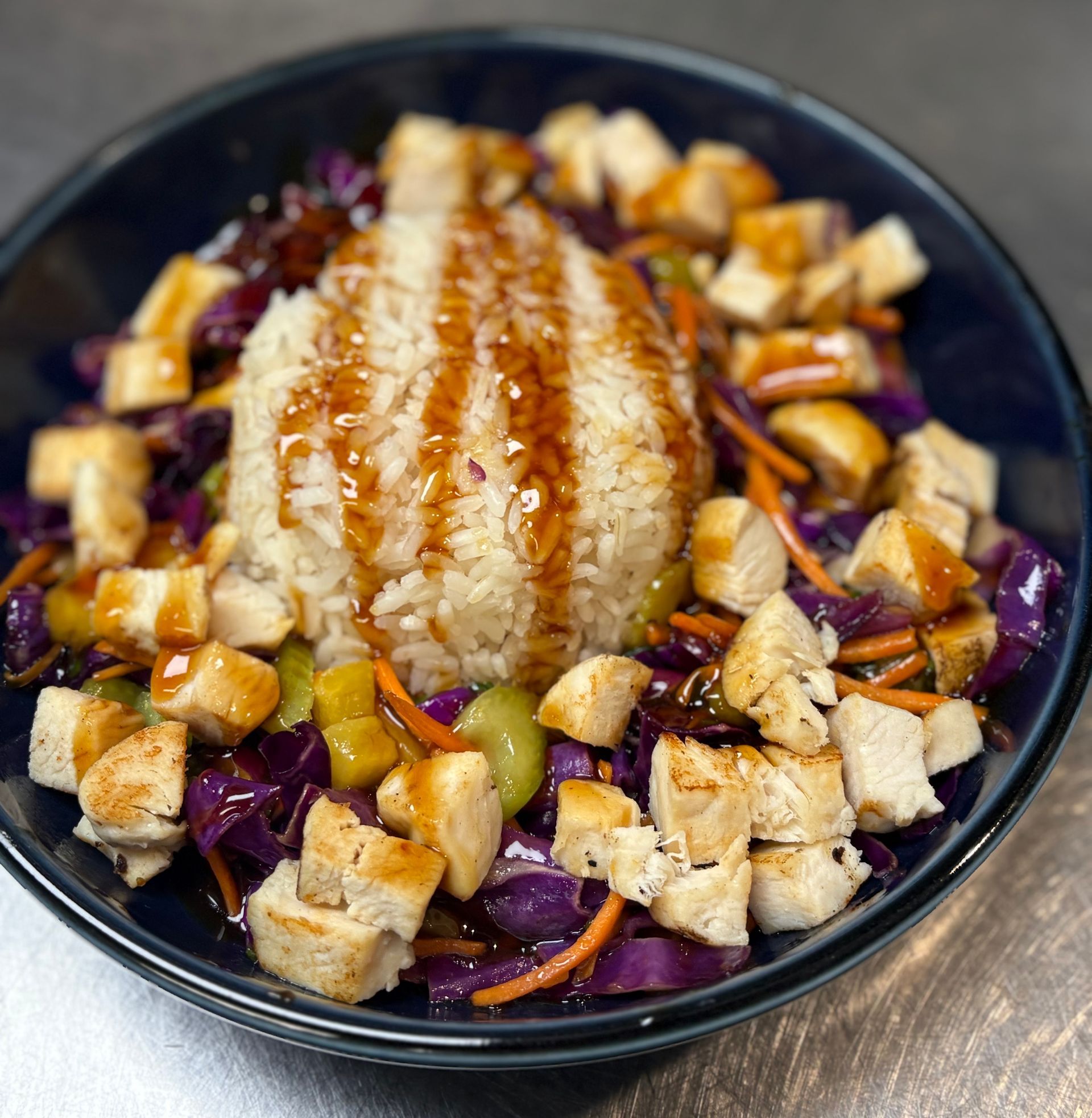 Chicken and rice bowl with vegetables and sauce in a dark blue bowl.