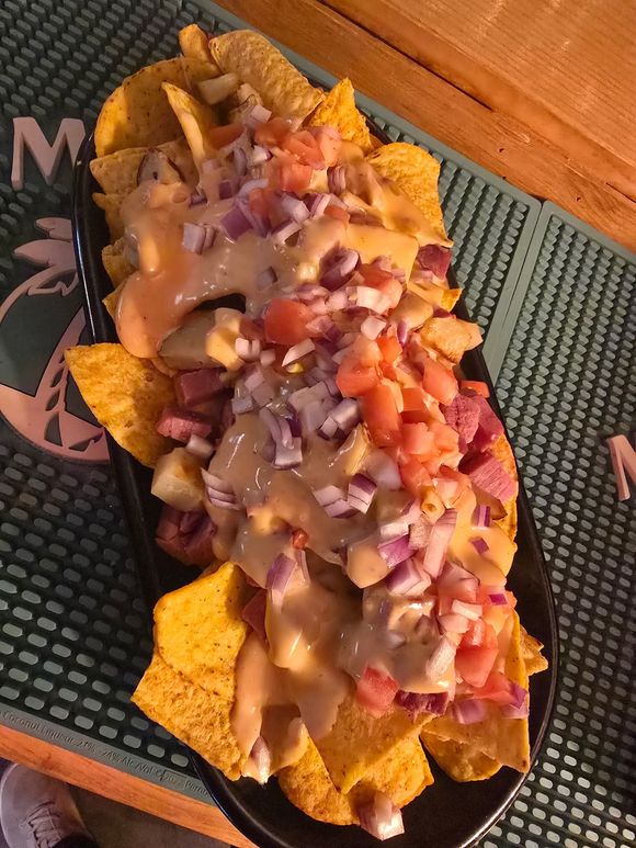 A plate of corn tortilla chips topped with melted cheese, diced tomatoes, red onions, and meat on a rubber bar mat.