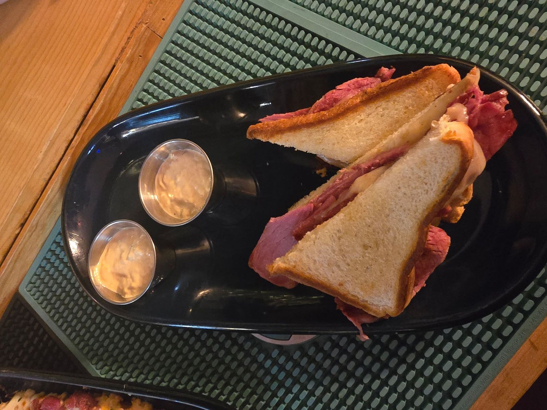 A halved meat sandwich with two small side portions of sauce on a black oval plate set on a green bar mat.