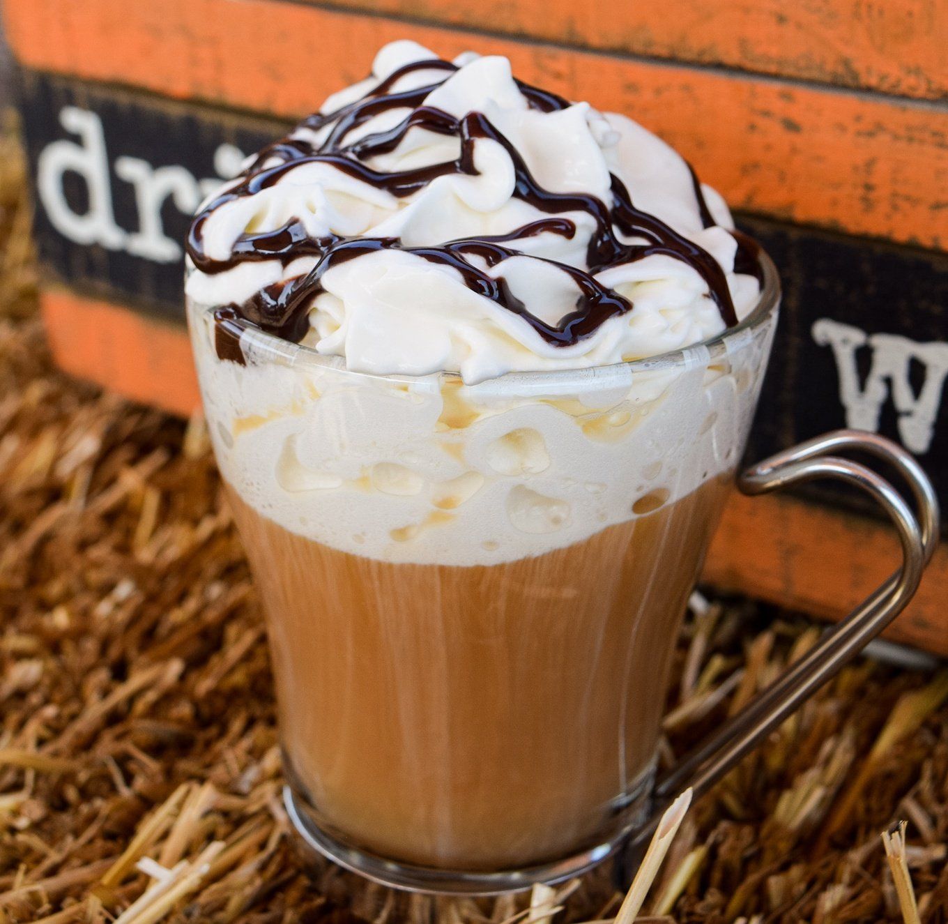 A clear glass mug with a coffee drink topped with whipped cream and chocolate syrup, sitting on straw.