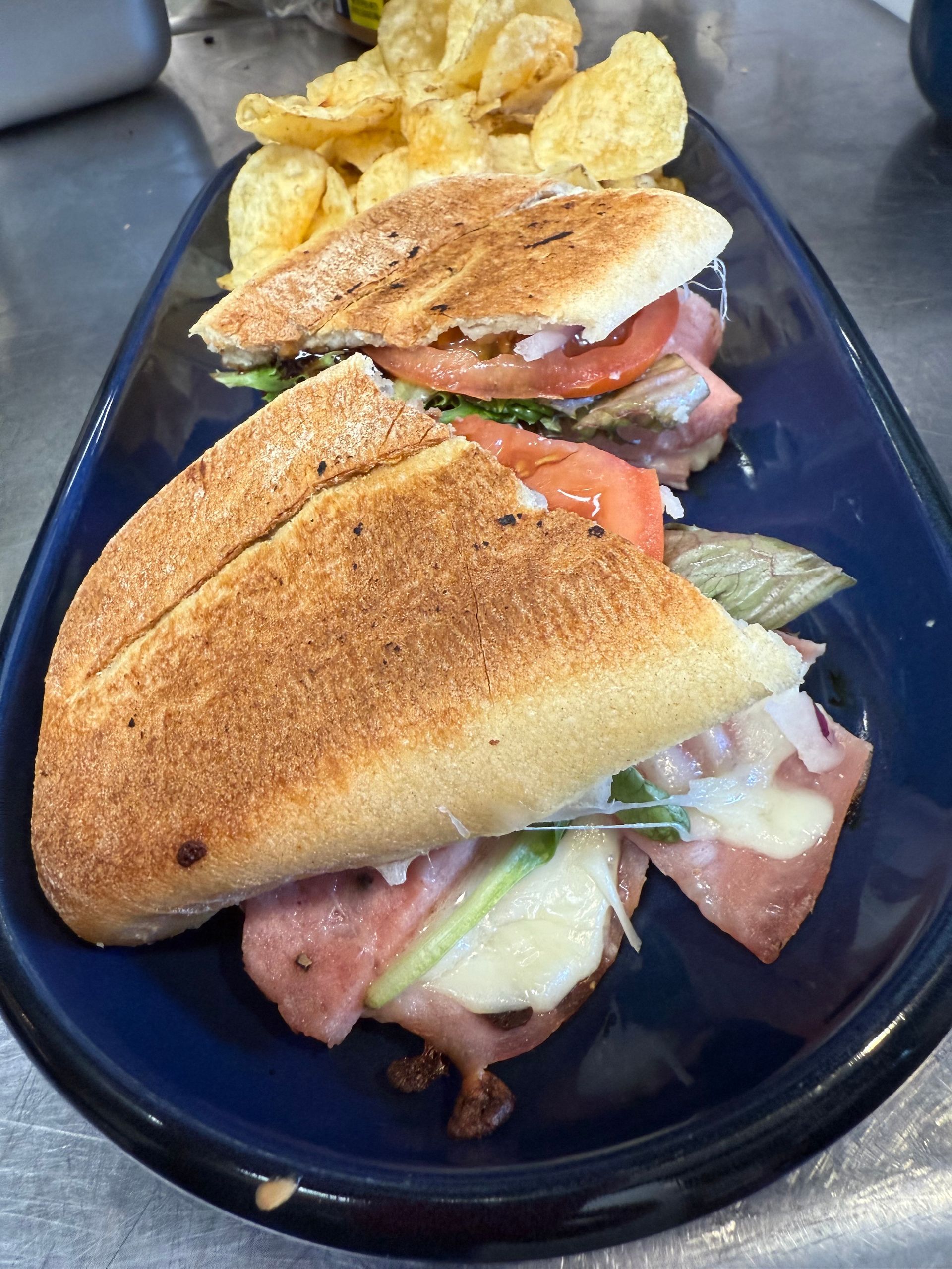 Two deli sandwiches on a blue plate with potato chips; sandwiches include ham, cheese, tomato, and greens.