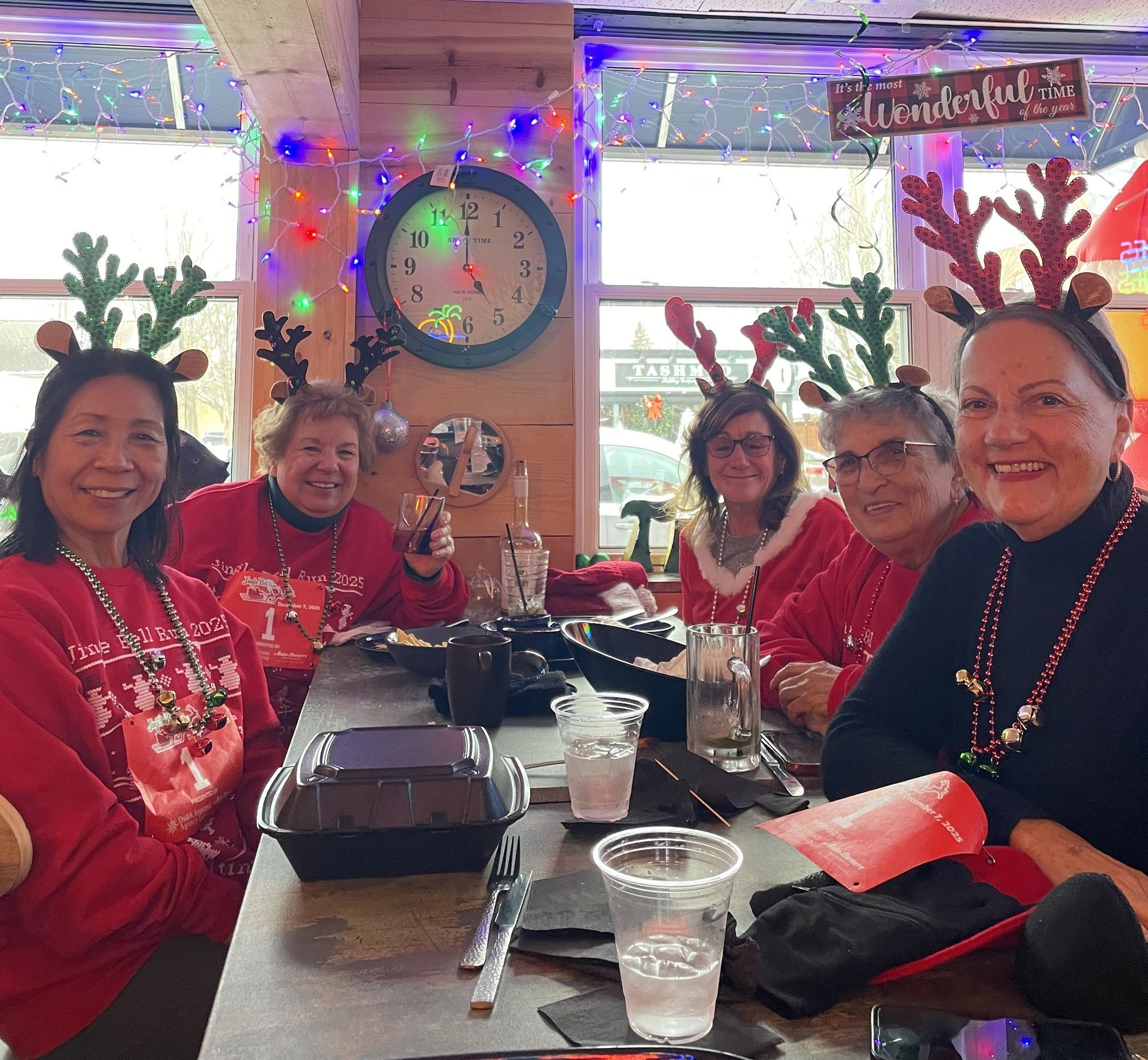 Five people in red sweaters and reindeer antlers sit at a decorated table, a festive restaurant setting.