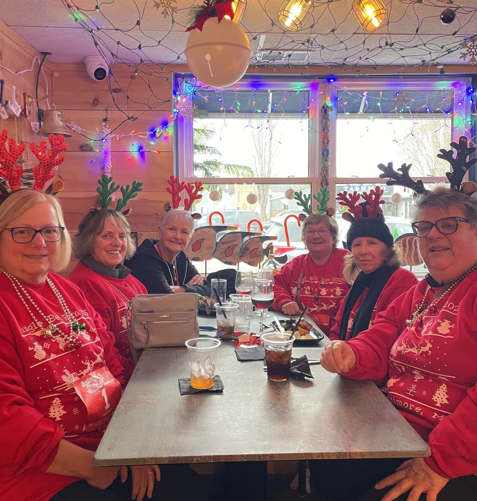 Six women in red sweaters and reindeer antlers at a restaurant table with Christmas decorations.