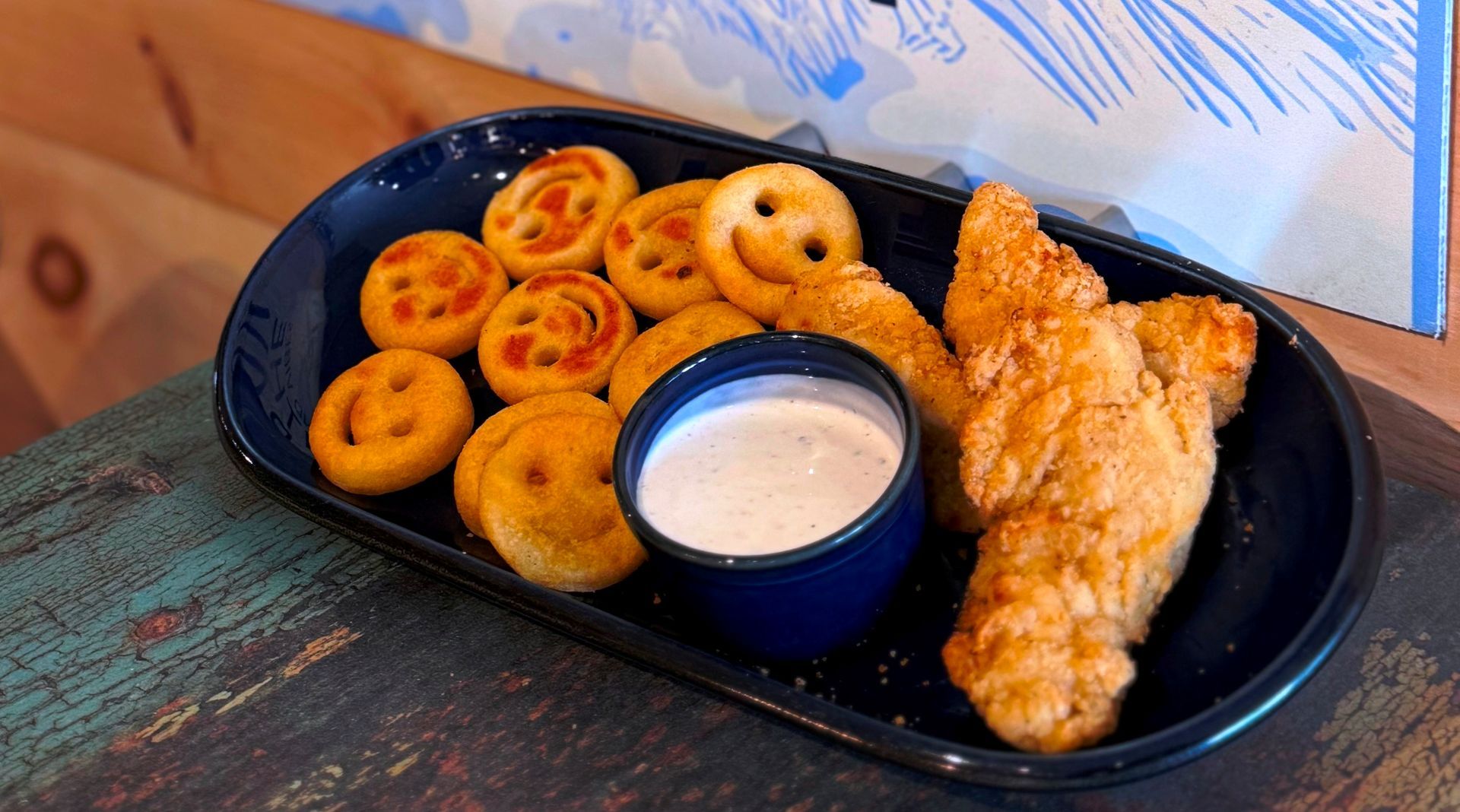 Plate of smiley-face potatoes, fried chicken strips, and dipping sauce on a blue dish.