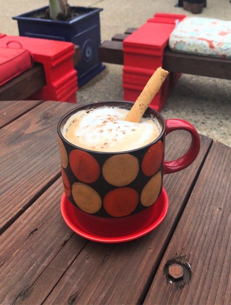 A coffee mug with orange and yellow polka dots, topped with foam and a cinnamon stick, sits on a red saucer on a wooden table.