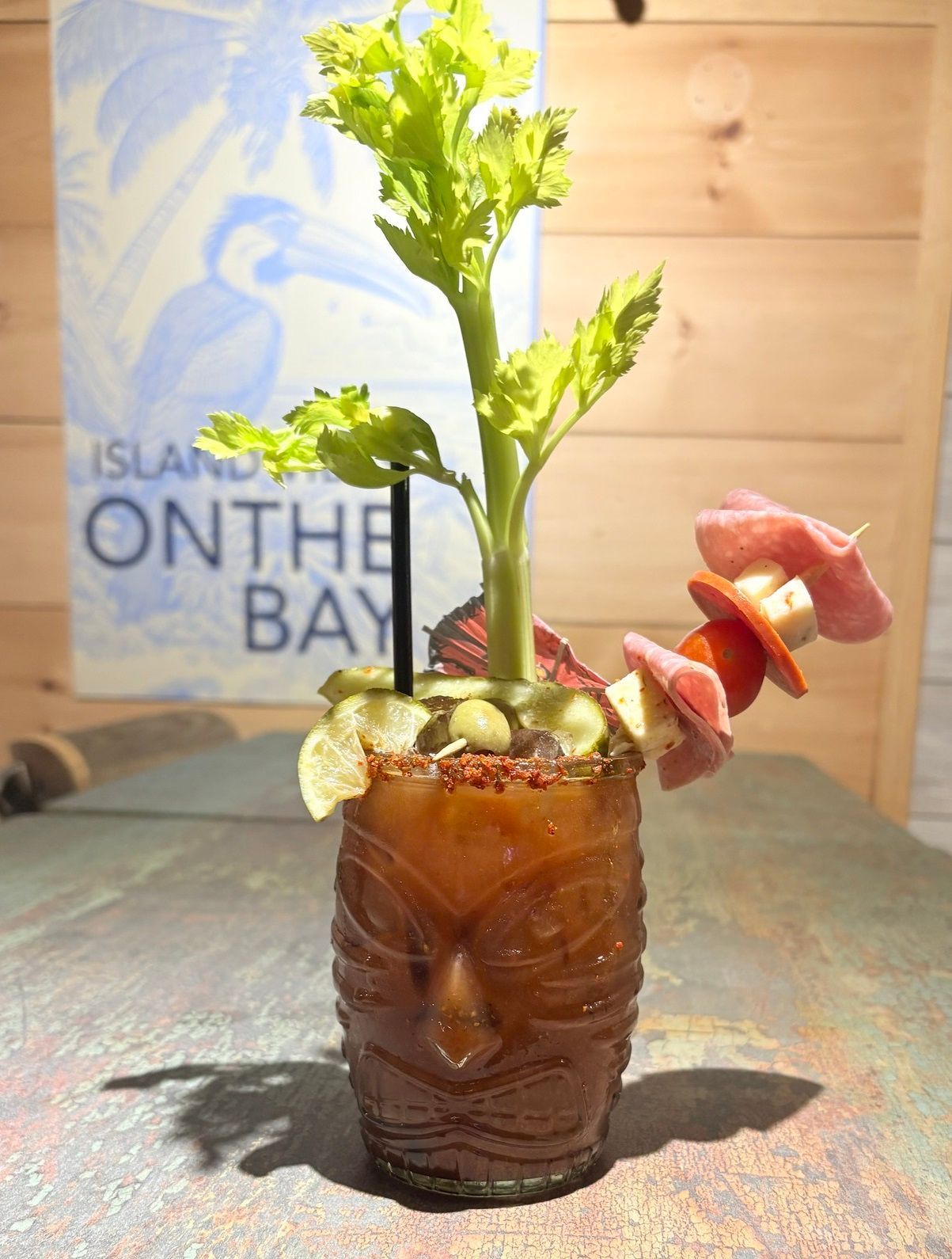 Tiki cocktail garnished with celery, meat, and tomato; set against a background.