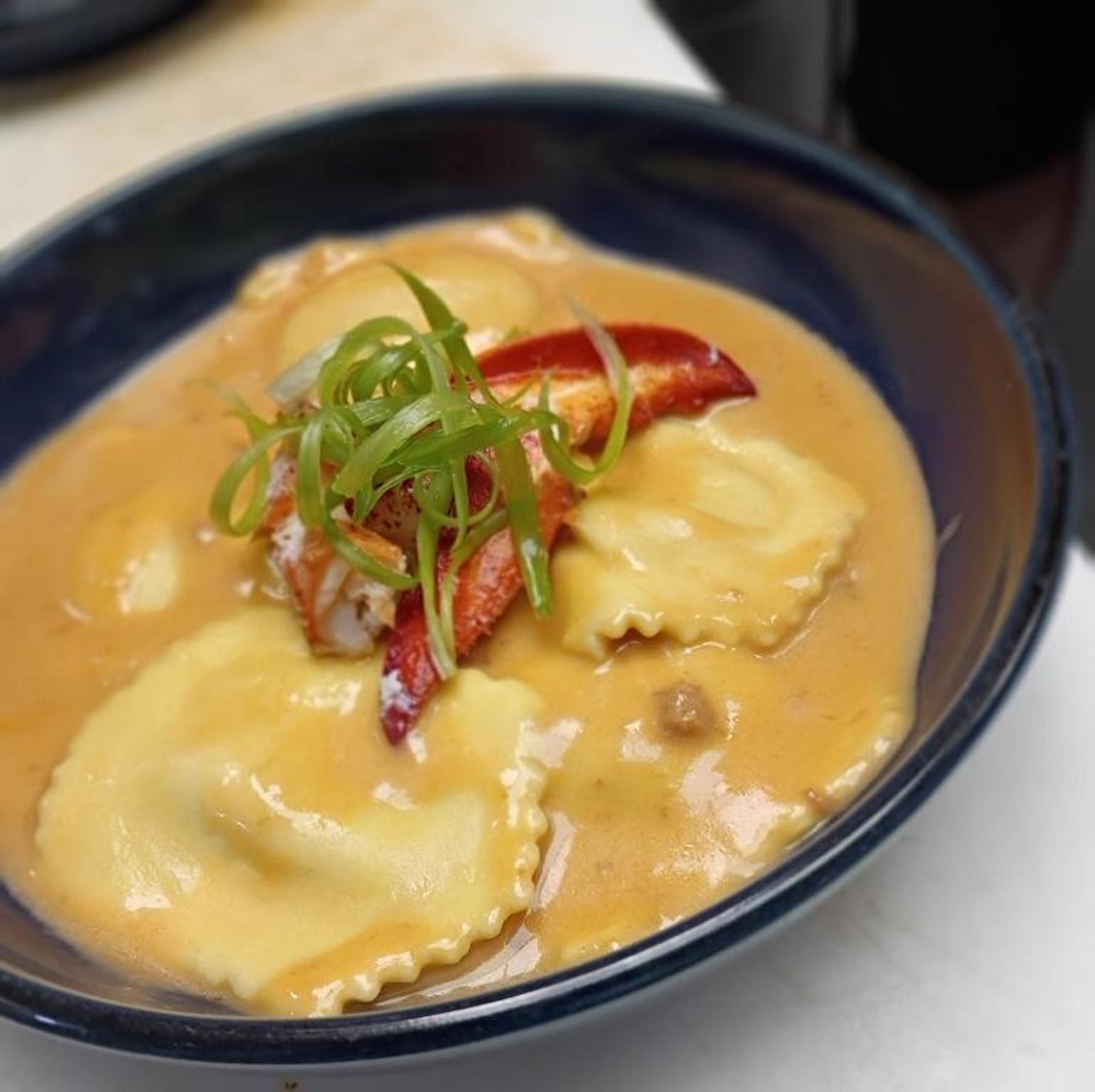 Lobster mac and cheese in black container, creamy sauce, lobster claw visible, sprinkled parsley, yellow cheese, wood background.