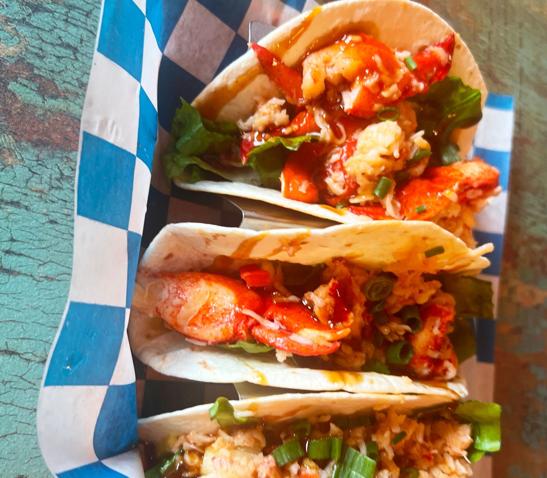 Three lobster tacos in tortillas with lettuce, sauce, and green onions, served in a blue and white checkered paper.