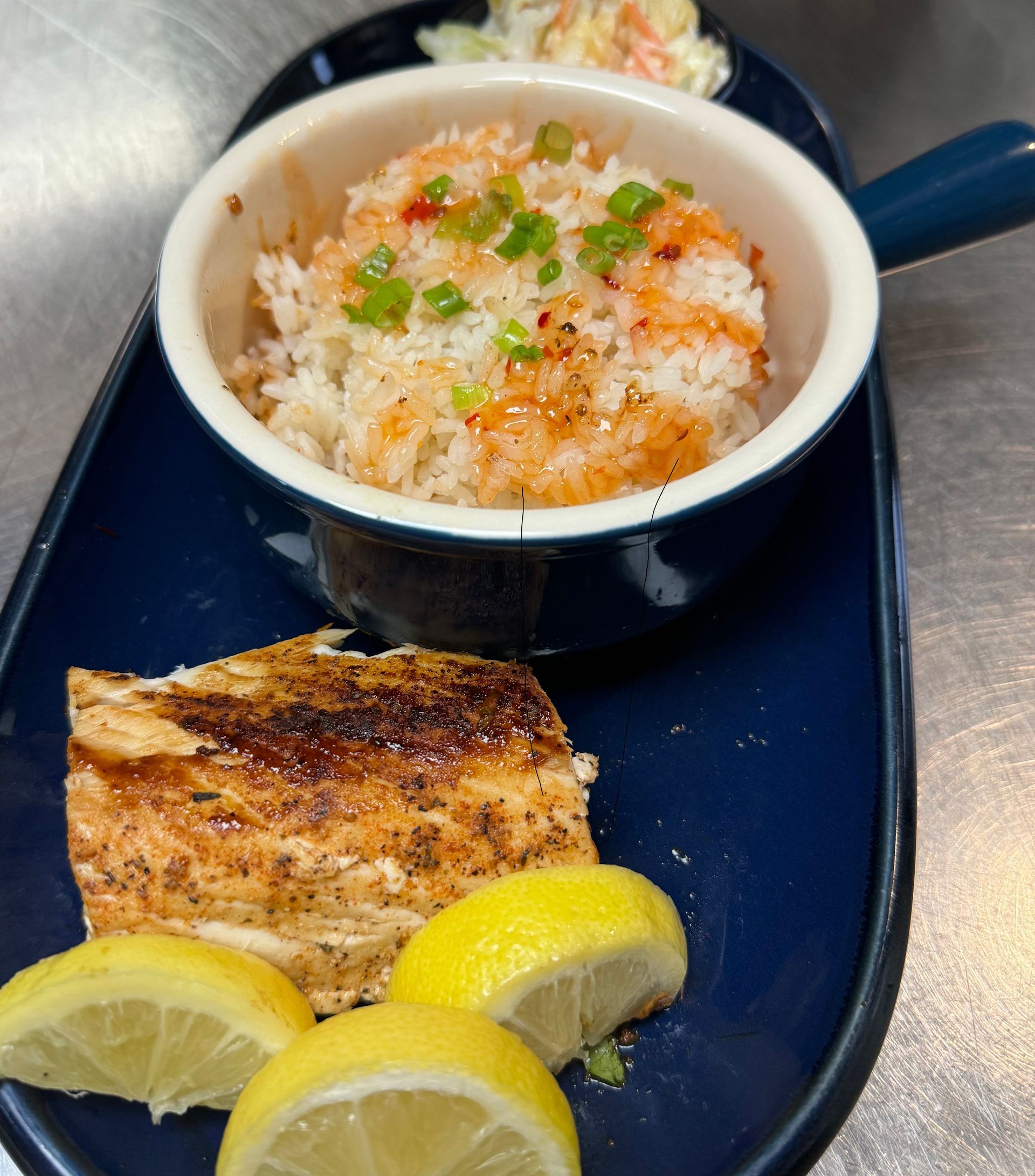 Grilled salmon with lemon slices, rice in a small dish, and coleslaw on a blue platter.