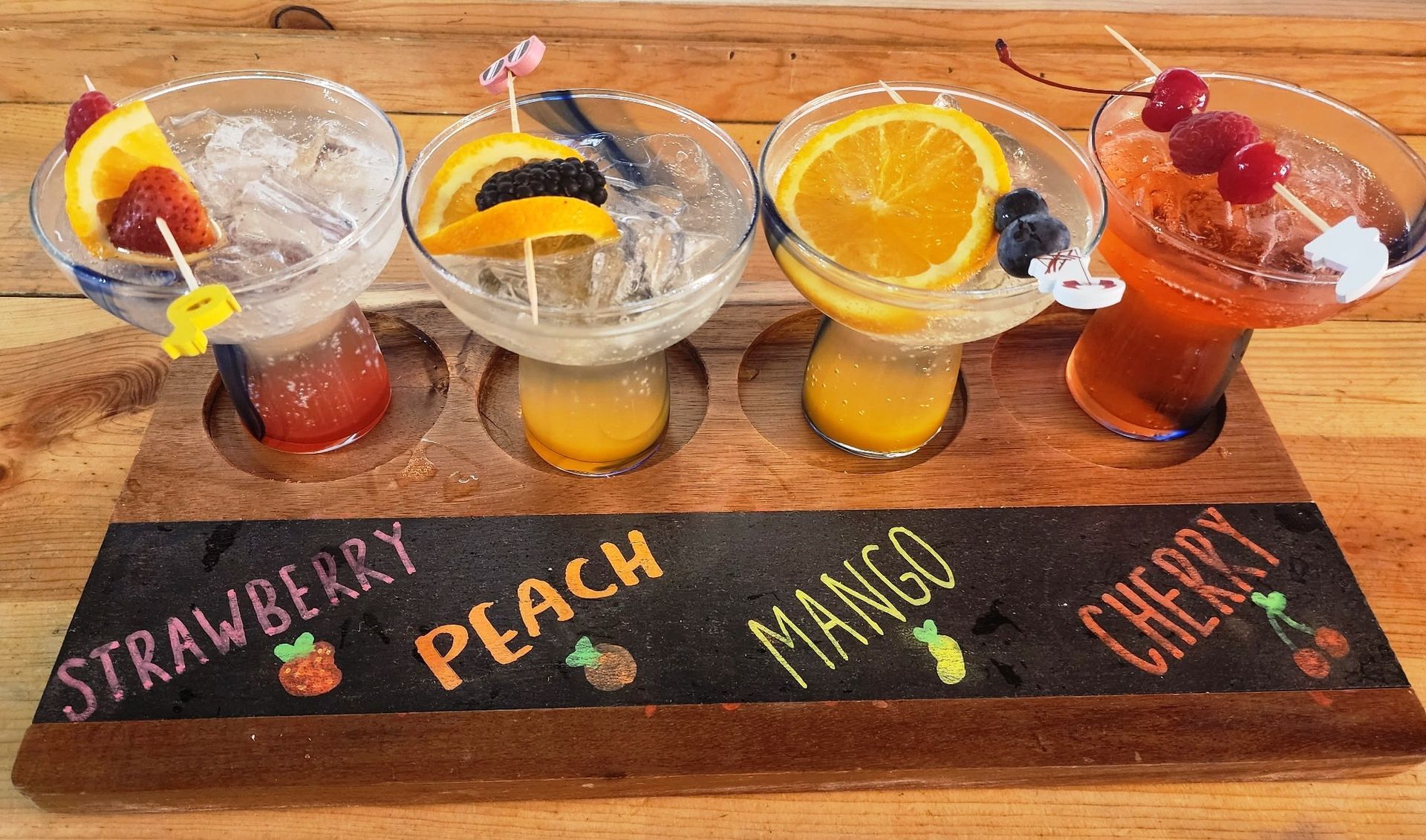 A flight of colorful cocktails: strawberry, peach, mango, and cherry, with fruit garnishes, on a wooden tray.