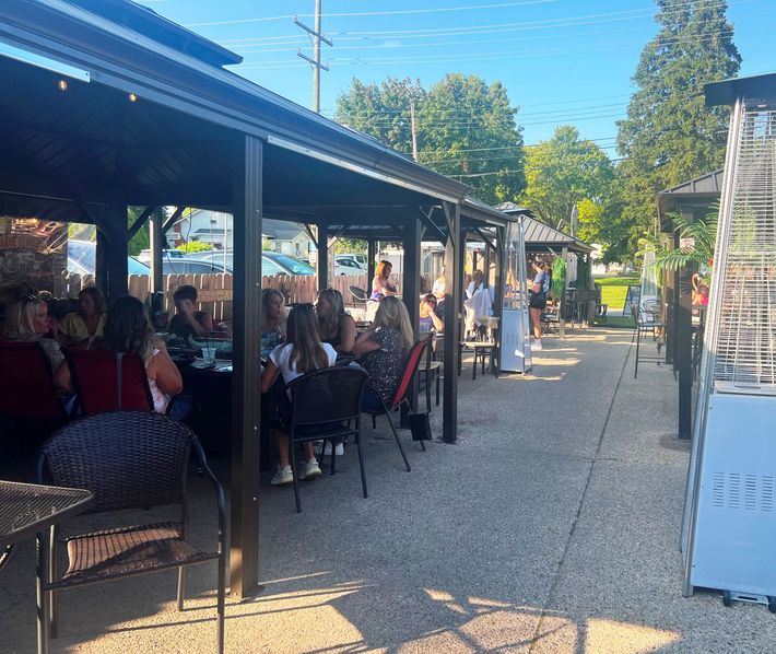 Outdoor restaurant seating area. People dining at tables under a canopy. Concrete patio, trees in background, sunny day.