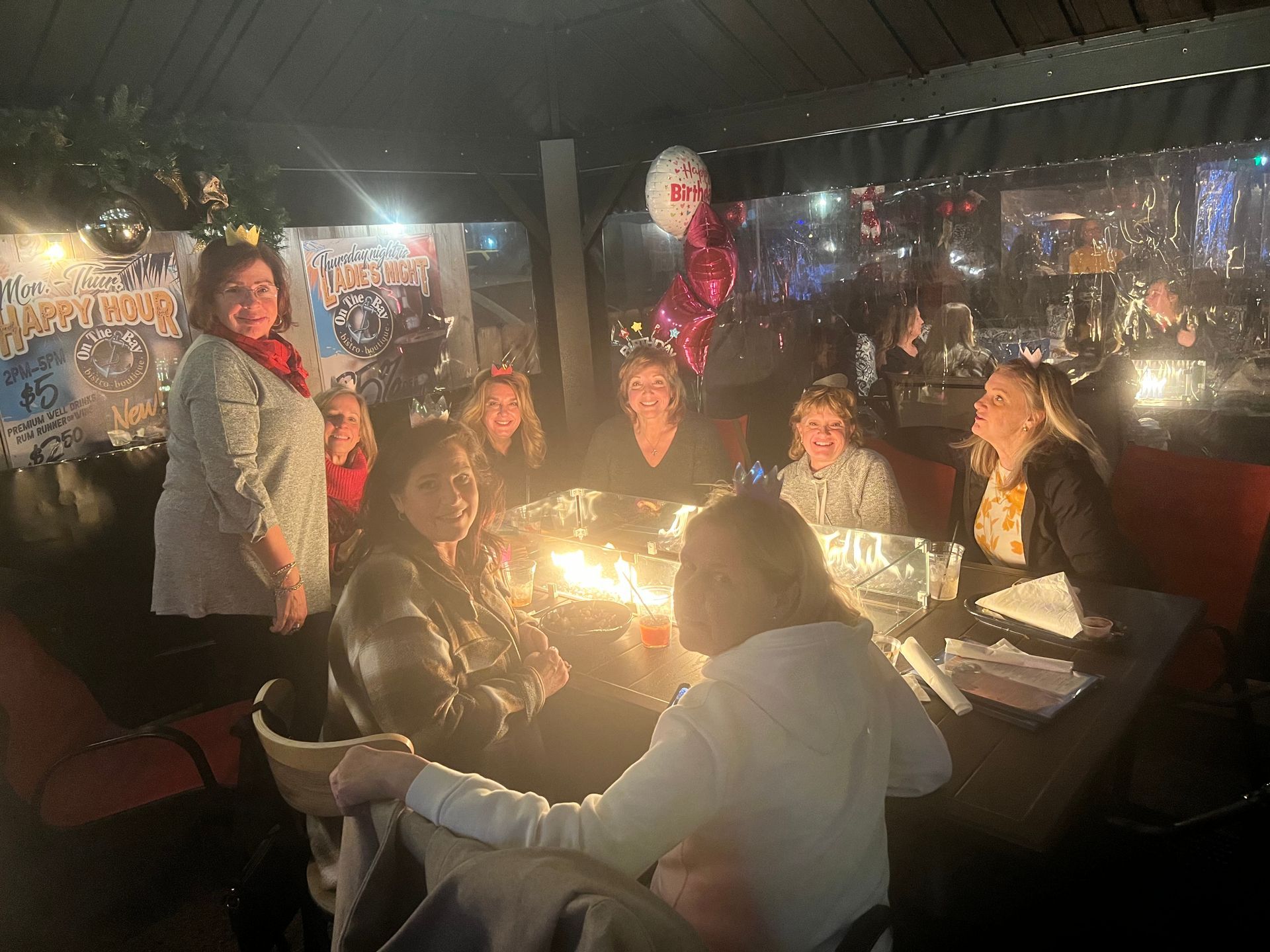 A group of people are sitting around a table with a fire pit in the middle at On The Bay restaurant, bar, and boutique in downtown New Baltimore Michigan
