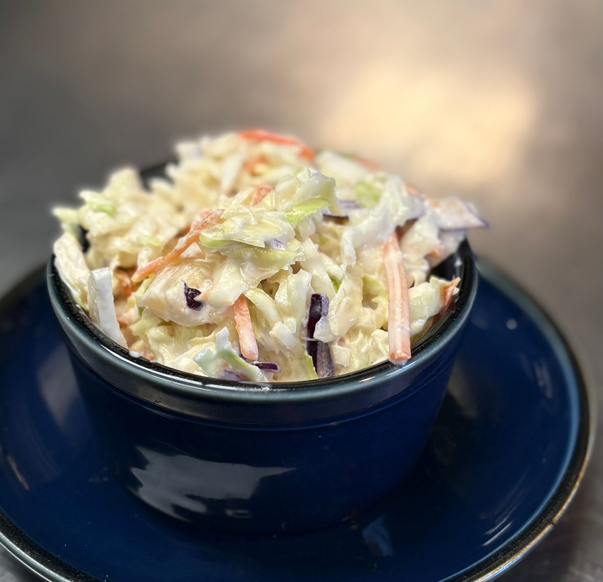 Coleslaw in a blue bowl on a blue saucer. Creamy dressing coats shredded cabbage and carrots.