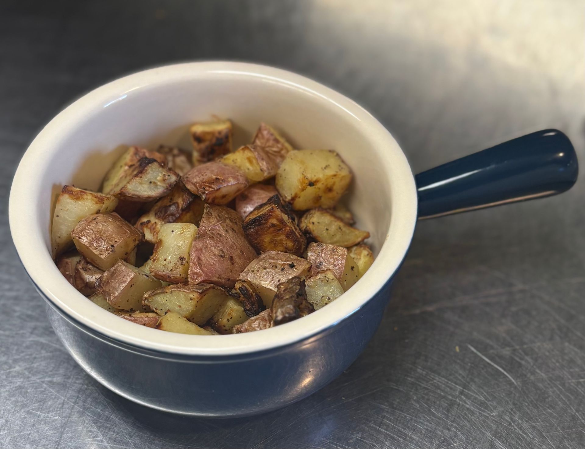 Roasted red potatoes in a small blue ceramic bowl with a handle, on a stainless steel surface.