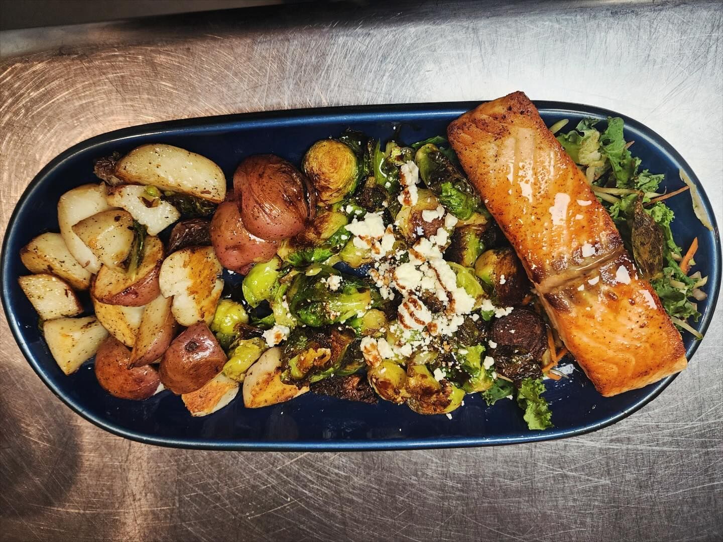 Plated salmon with roasted potatoes, Brussels sprouts, and greens on a dark oval dish