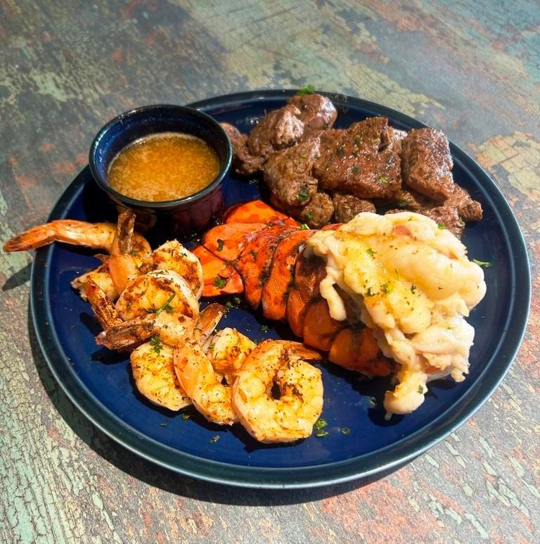 Surf and turf plate: grilled shrimp, lobster tail, steak, and butter sauce on a blue plate.