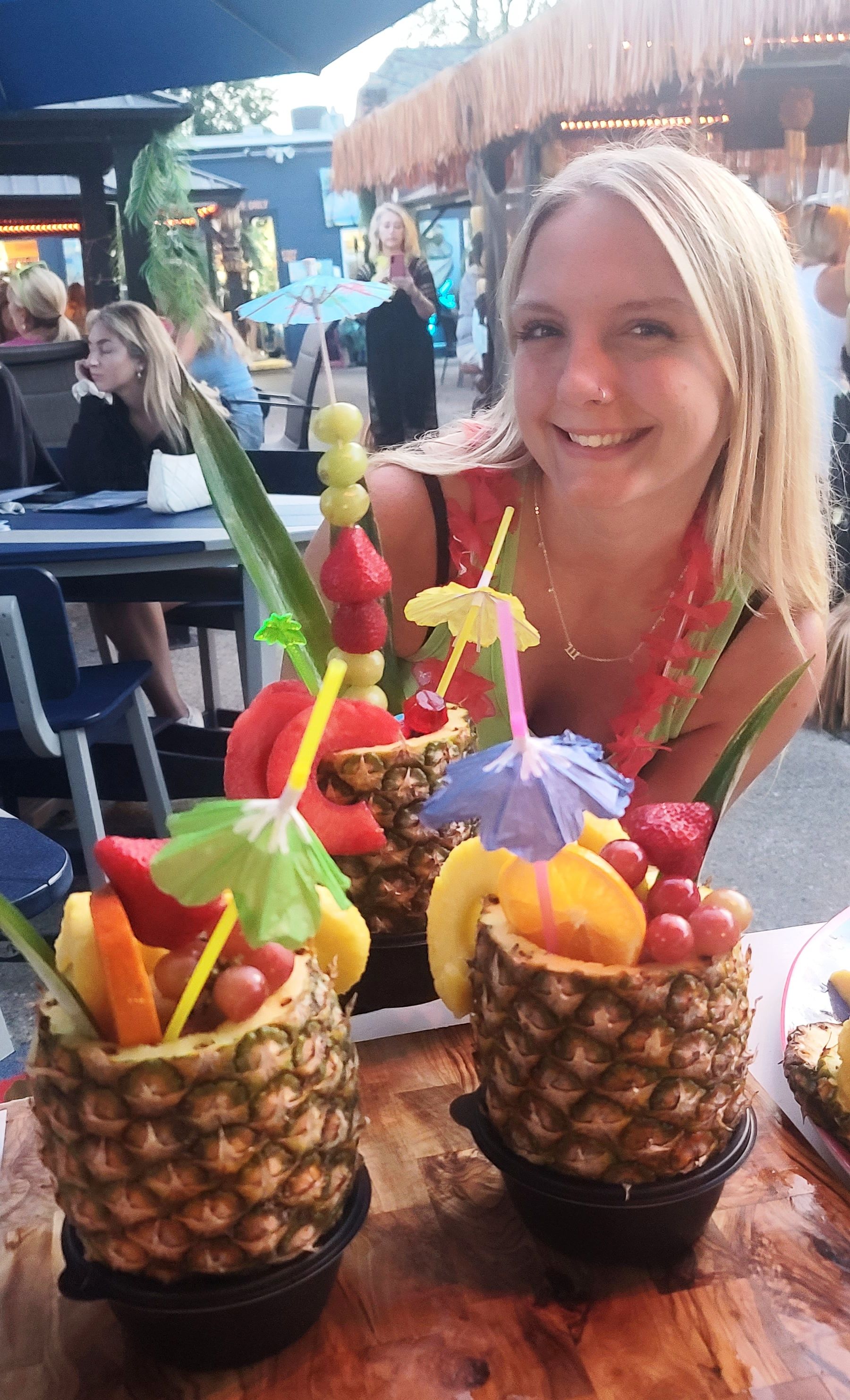 Blonde woman smiles, stands behind fruit cocktails served in pineapples at an outdoor tiki bar.