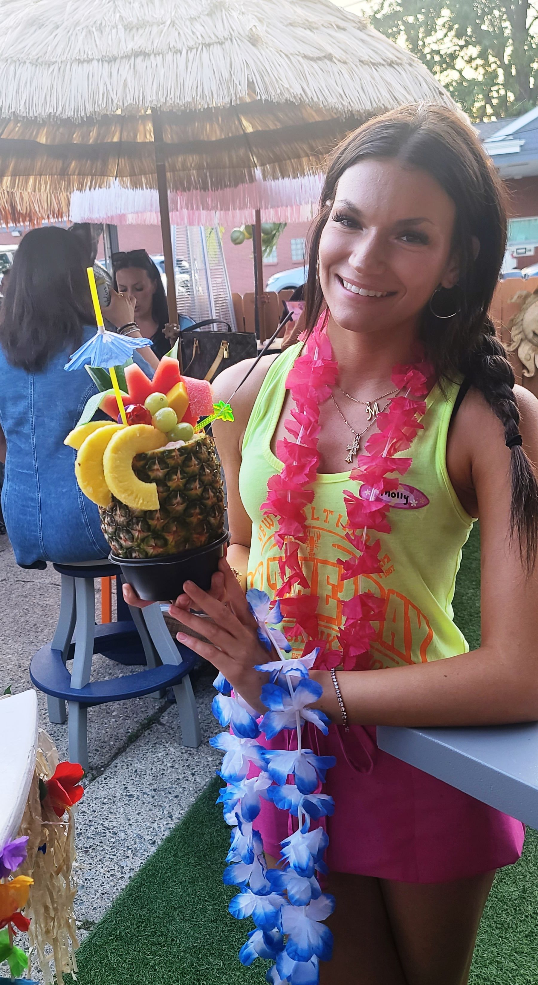 Woman in tropical attire smiling, holding a pineapple cocktail in an outdoor tiki bar setting.
