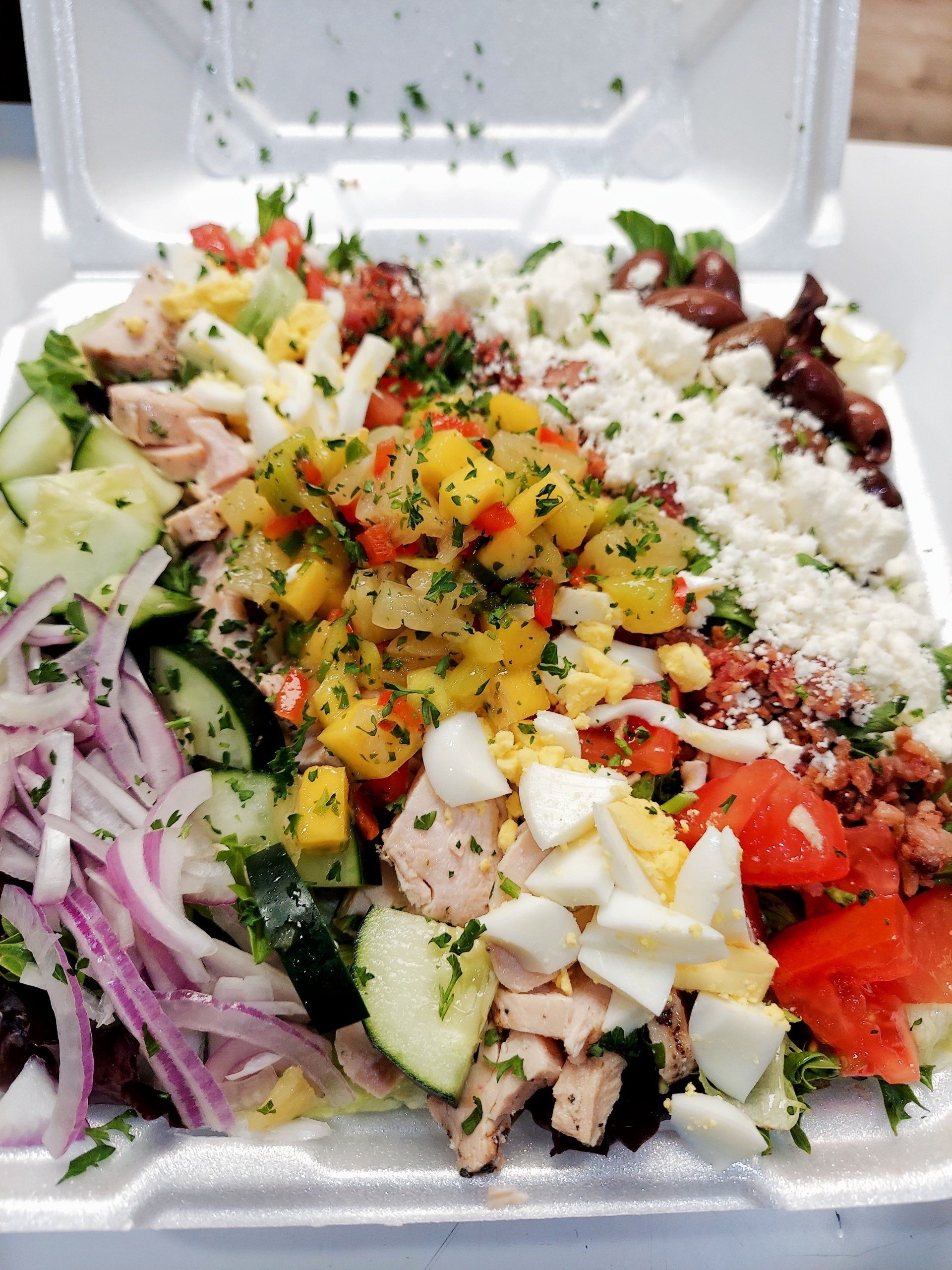Salad in a white container, with various colorful toppings like eggs, tomatoes, and feta cheese.