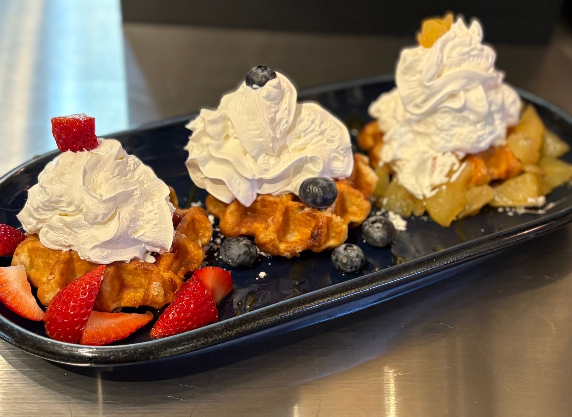 Three waffles with whipped cream and fruit on a blue rectangular plate. Strawberries, blueberries, and peaches.