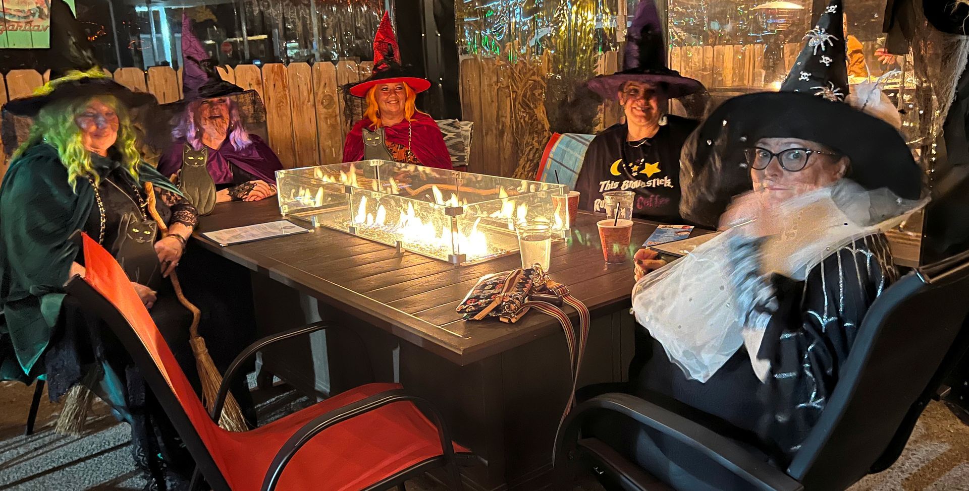 Five people in witch hats around a table with fire feature, outdoors.