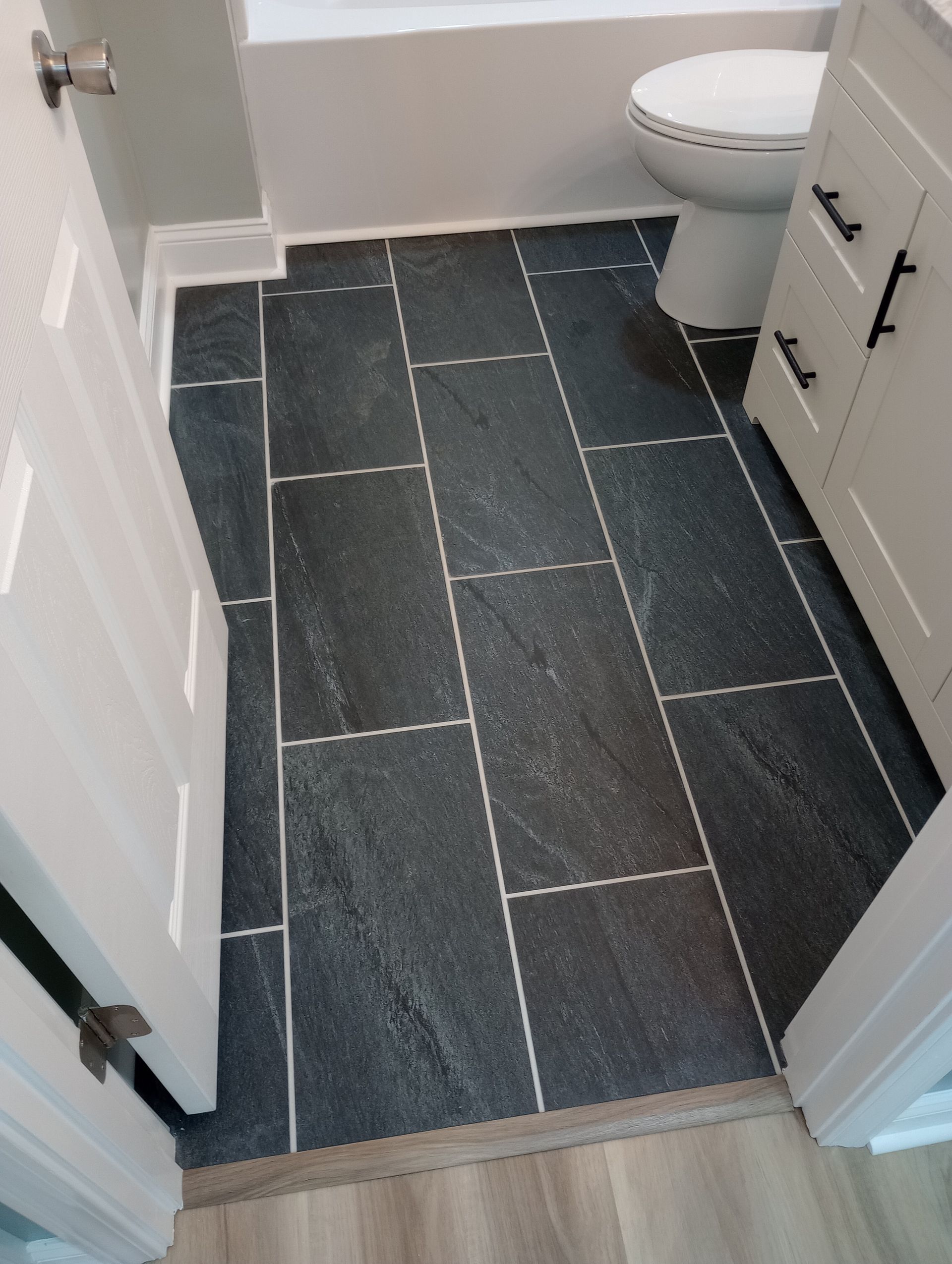 Dark slate-tiled bathroom floor with white grout, doorway, toilet, and vanity.